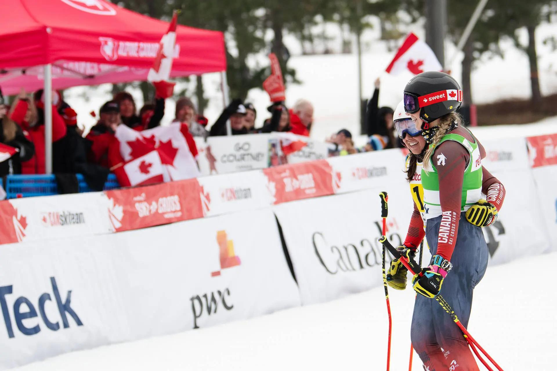 two female skiers together in front supporters waving Canadian flags